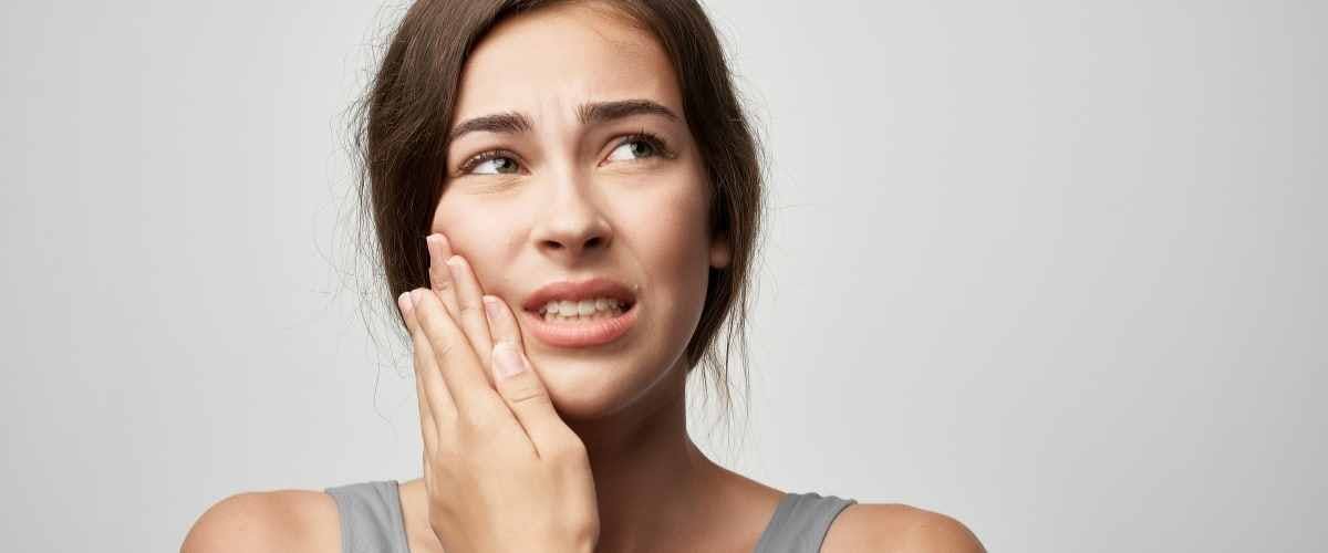 A woman with brown hair in a gray tank top holds her cheek with a pained expression, suggesting a toothache or jaw discomfort against a plain background.